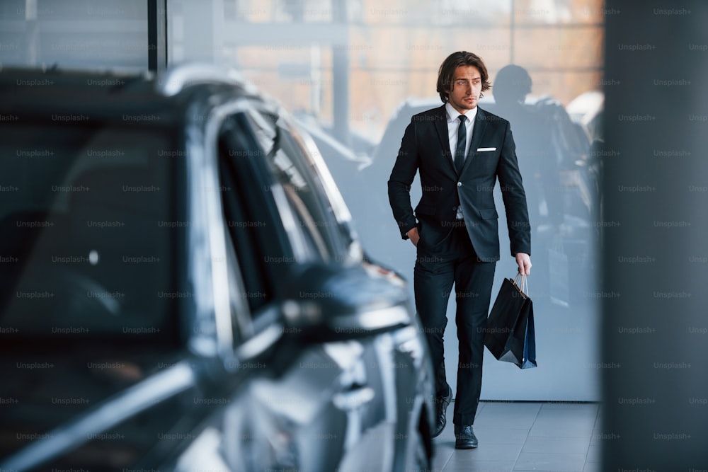 Retrato de un joven y guapo hombre de negocios con traje negro y corbata y con bolsa de compras cerca del automóvil moderno.