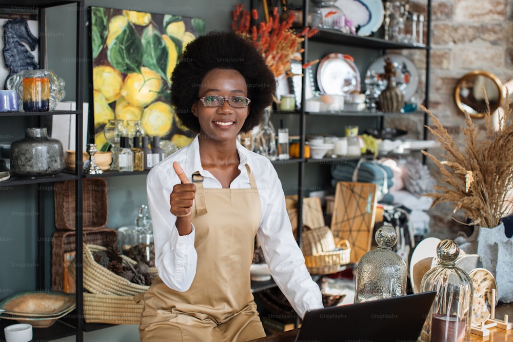 Beautiful afro american woman showing thumb up and smiling on camera while working at decor shop with laptop. Concept of people, technology and sale.