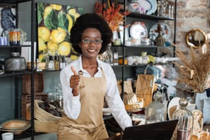 Beautiful afro american woman showing thumb up and smiling on camera while working at decor shop with laptop. Concept of people, technology and sale.