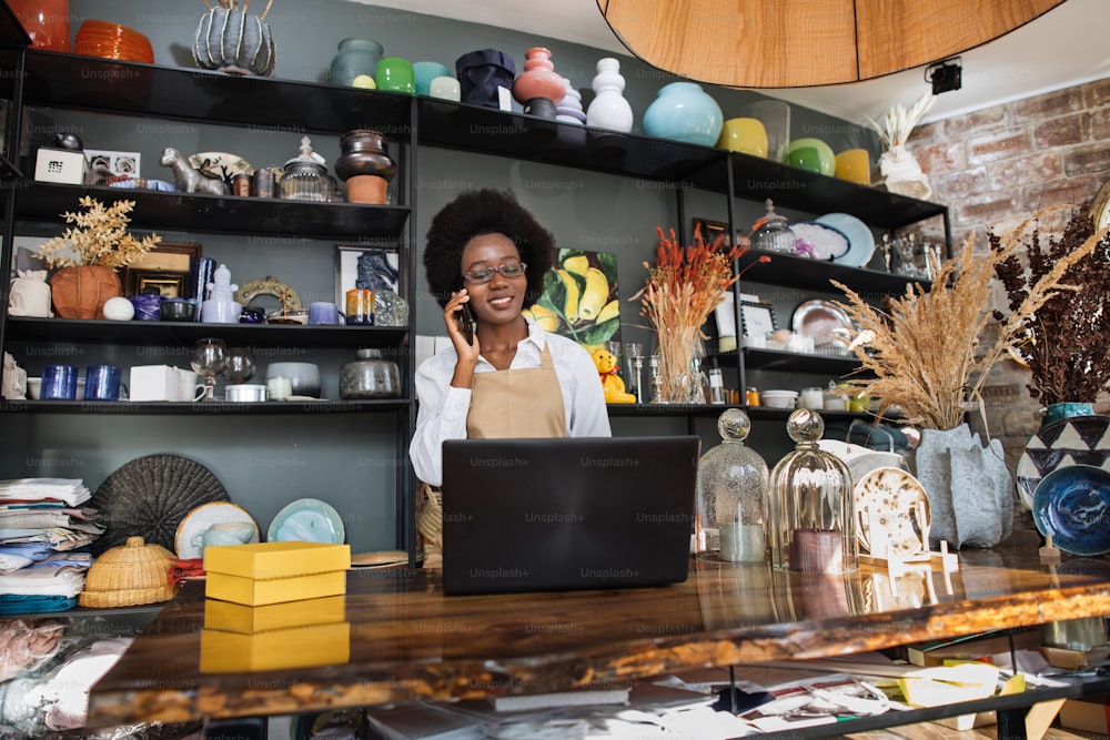 Afro saleswoman talking on mobile and typing on laptop at decor shop. Various beautiful decor lying on shelves. Modern gadgets for work.