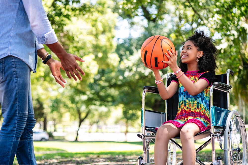 A little girl in a wheelchair having fun with her father while playing basketball together at park.