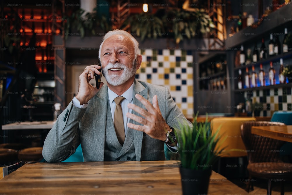 Feliz e bonito empresário sênior sentado no restaurante e esperando pelo almoço. Ele está usando smartphone e conversando com alguém. Conceito de estilo de vida para idosos de negócios.