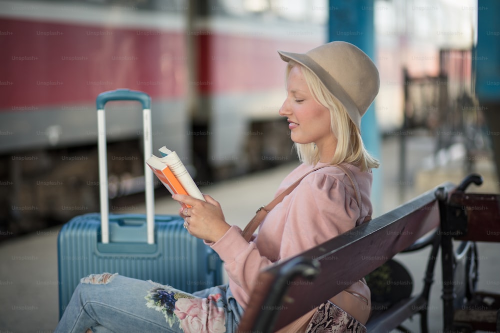 Young woman sitting on bench, waiting for a train and reading book.