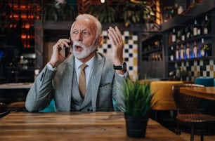 Happy and handsome senior businessman sitting in restaurant and waiting for lunch. He is using smart phone and talking with someone. Business seniors lifestyle concept.