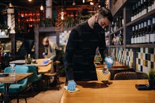 Junge Restaurantangestellte reinigen und desinfizieren Tische und Oberflächen gegen die Coronavirus-Pandemie. Sie tragen Gesichtsschutzmasken und Handschuhe.