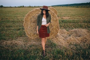 Stylish woman in hat standing at hay bale in summer evening in field. Atmospheric tranquil moment. Young fashionable female relaxing at haystack,  vacation in countryside
