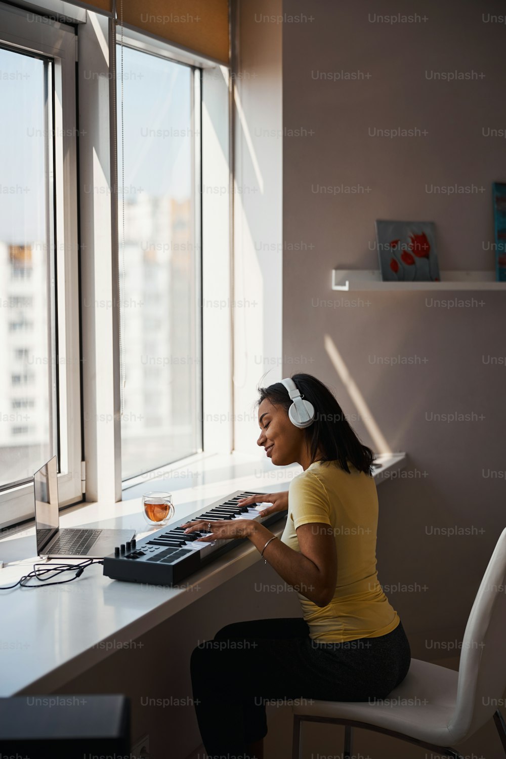 Hermosa músico femenina con auriculares sentada en el alféizar de la ventana y sonriendo mientras toca la melodía en el sintetizador y usa la computadora portátil