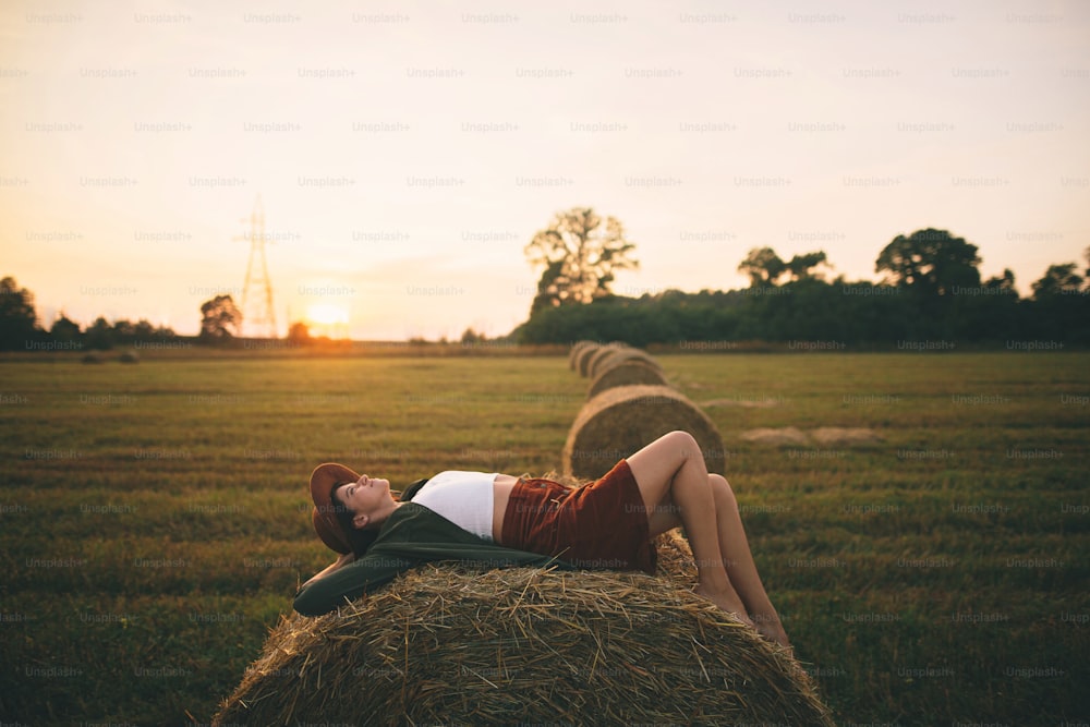 Schöne sorglose Frau mit Hut, die im Sonnenuntergangslicht auf dem Heuhaufen liegt und den Abend im Sommerfeld genießt. Junges, glückliches Weibchen entspannt sich auf Heuballen auf dem Land. Atmosphärischer ruhiger Moment
