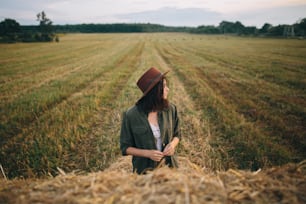 Beautiful stylish woman in hat standing at hay bale in summer evening field. Young fashionable female relaxing at haystacks, summer vacation in countryside. Tranquility
