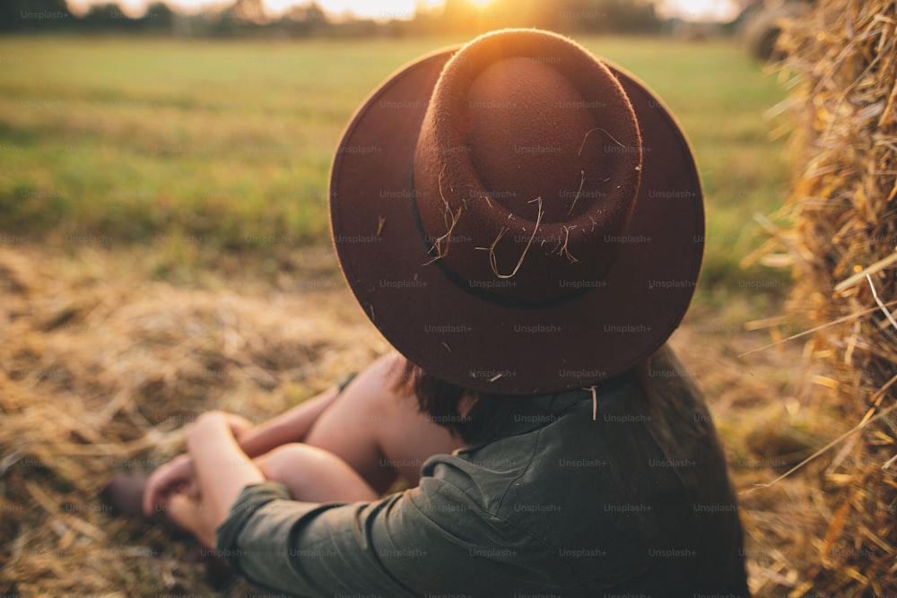 Stylish woman in hat looking at sunset light, sitting at haystacks in summer field. Atmospheric tranquil moment in countryside. Young female enjoying evening at hay bale in warm sunshine