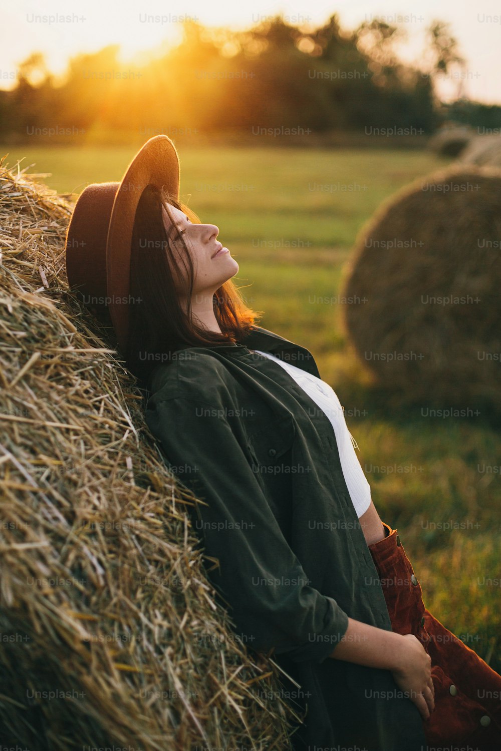 Beautiful stylish woman in hat relaxing on haystacks in sunset light in summer field. Atmospheric tranquil moment in countryside. Young female enjoying evening at hay bale in warm sunshine