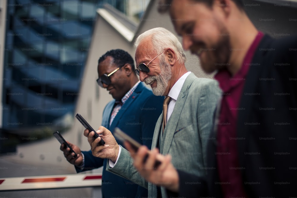 Business people typing messages.  Three business men in the city standing on street and using mobile phones.  Focus is on background.