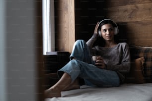 Resting with joy. Beautiful young woman listening to music while sitting on sofa at home near the window