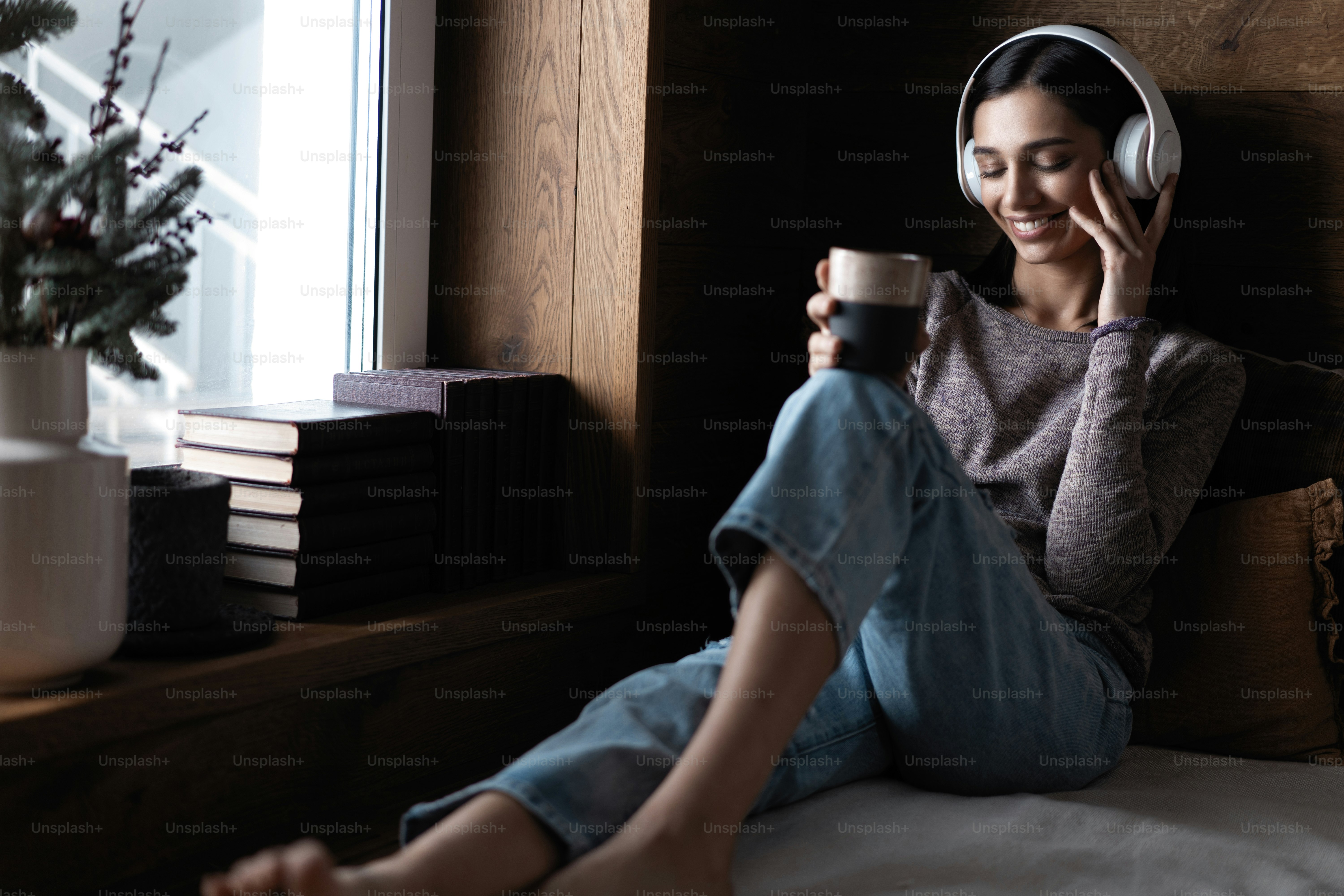 Beristirahat dengan sukacita. Wanita muda cantik mendengarkan musik sambil duduk di sofa di rumah dekat jendela