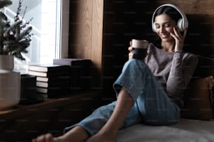 Resting with joy. Beautiful young woman listening to music while sitting on sofa at home near the window