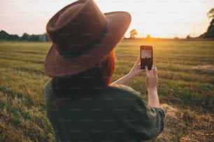 Femme élégante prenant une photo du coucher de soleil sur le téléphone dans le champ d’été. Jeune femme tenant un smartphone et capturant le soleil chaud du soir dans la campagne. Beau moment atmosphérique