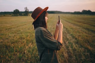 Hermosa mujer elegante con sombrero tomando foto de la puesta de sol en el teléfono en el campo de verano. Retrato de una mujer joven y atractiva que sostiene un teléfono inteligente y captura la noche bajo un sol cálido. Momento atmosférico