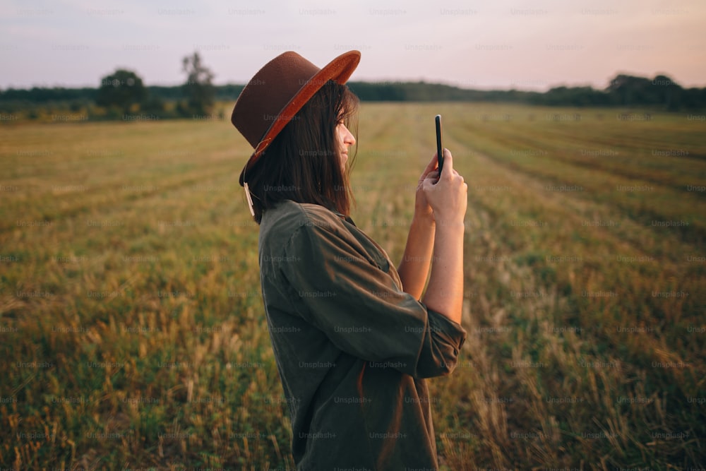 Beautiful stylish woman in hat taking photo of sunset on phone in summer field. Portrait of young attractive female holding smartphone and capturing evening in warm sunshine. Atmospheric moment