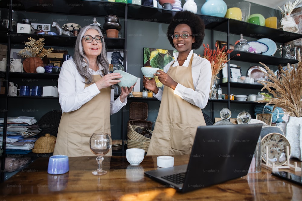 Caucasian senior woman and african young woman working together at modern shop with various decor, doing inventory, holding original ceramic cups and smiling at camera