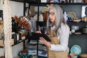 Charming aged woman using digital tablet while doing inventory at decor shop. Competent saleswoman with grey hair wearing eyeglasses, white shirt and beige apron.