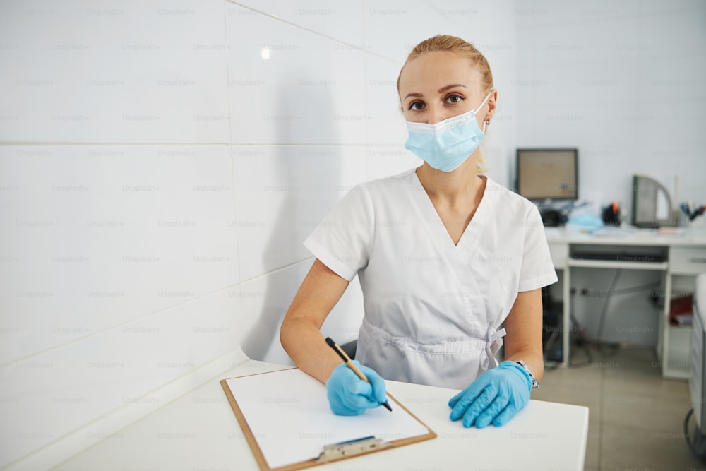 Junge Frau in medizinischer Kleidung blickt nach vorne, während sie eine Maske im Gesicht mit einem Stift in der rechten Hand über dem Notizbuch trägt