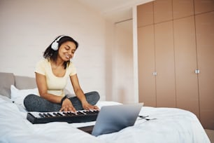 Hermosa músico femenina sentada en la cama y sonriendo mientras toca la melodía en un instrumento musical electrónico