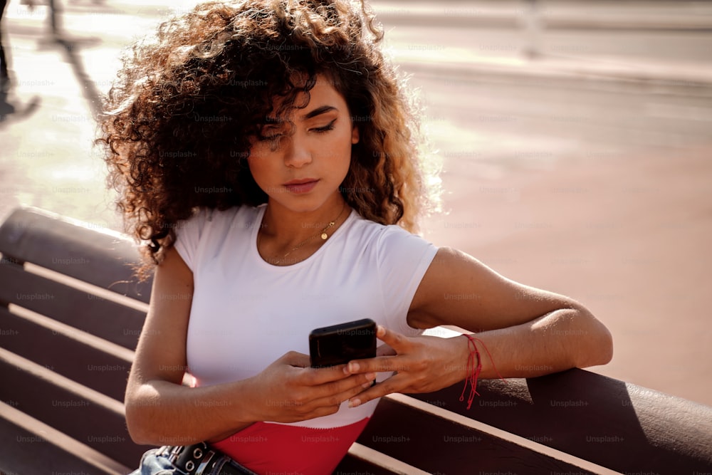 Cheerful colombian woman using mobile phone, reading something on social media. Mixed race girl with afro hair holding smartphone. Outdoor photo.