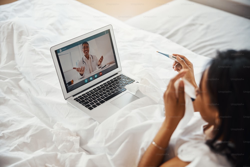 Gros plan d’une femme grippée tenant un thermomètre numérique et utilisant un ordinateur portable tout en parlant avec un médecin par appel vidéo