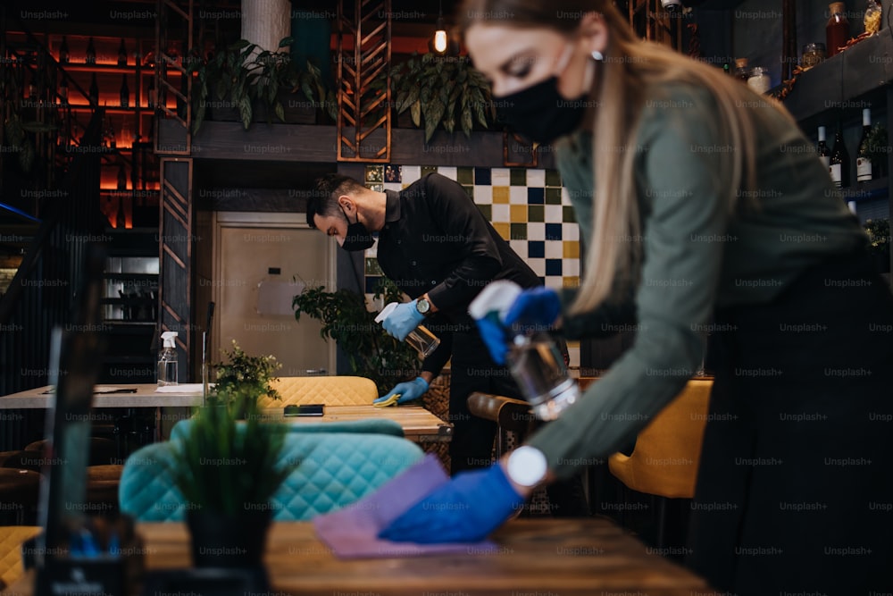 Giovani camerieri di ristoranti che puliscono e disinfettano tavoli e superfici contro la pandemia di Coronavirus. Indossano maschere protettive e guanti.