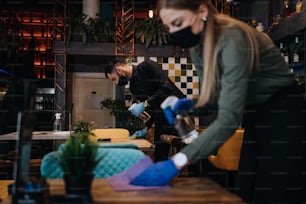 Young restaurant workers waiters cleaning and disinfecting tables and surfaces against Coronavirus pandemic disease. They are wearing protective face masks and gloves.