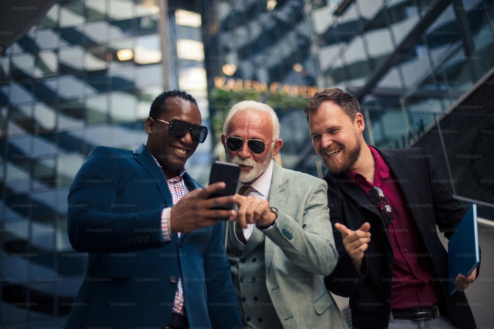 Colegas de negocios leyendo un mensaje de texto en un teléfono inteligente.  Tres hombres de negocios en la ciudad parados en la calle y usando el teléfono móvil.