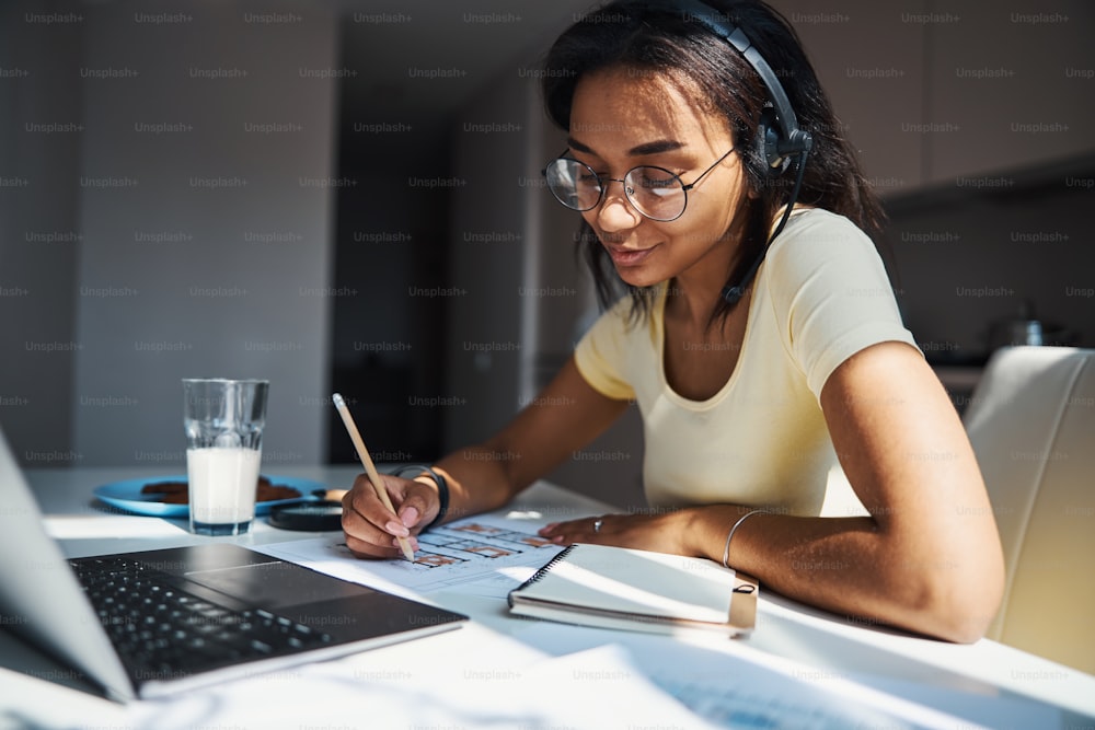 Charming female architect in glasses sitting at the table with laptop and studying building plan while working on project