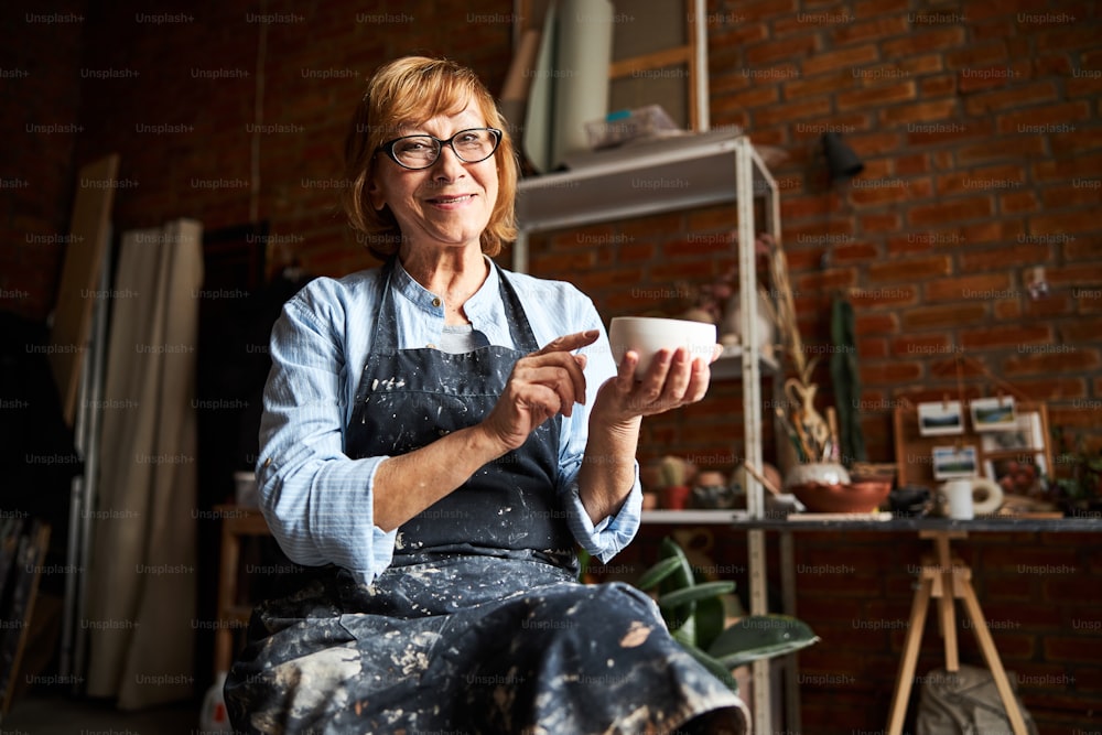 Mujer mayor alegre con delantal mirando a la cámara y sonriendo mientras sostiene un cuenco de barro hecho a mano