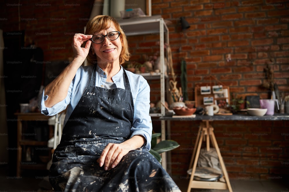 Nice female potter in apron looking at camera and smiling while adjusting glasses