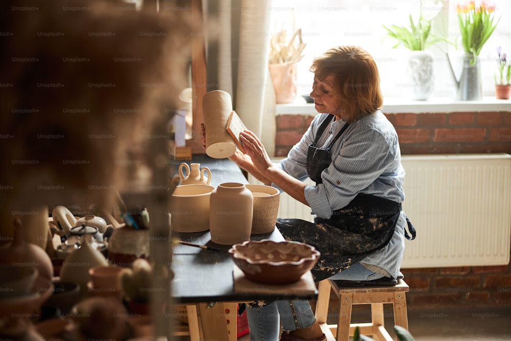 Nette alte Frau in Schürze polierende Tonvase mit Schleifblock beim Sitzen am Tisch mit handgemachter Keramik