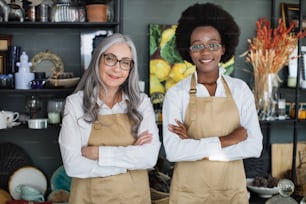 Front view of two multiracial saleswomen in beige aprons and eyeglasses smiling and posing with arms crossed at decor store. Concept of sale and service.