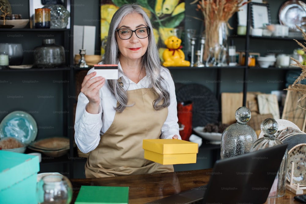 Close up of experienced mature saleswoman holding credit card in one hand and yellow paper box in another. Concept of payment, purchase and discount.