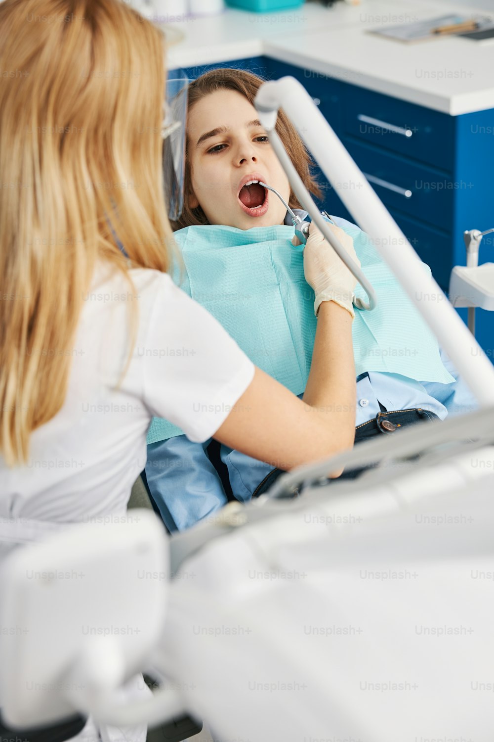Stomatologist in facial shield spraying teeth in opened mouth of a kid with dental pneumatic spray gun