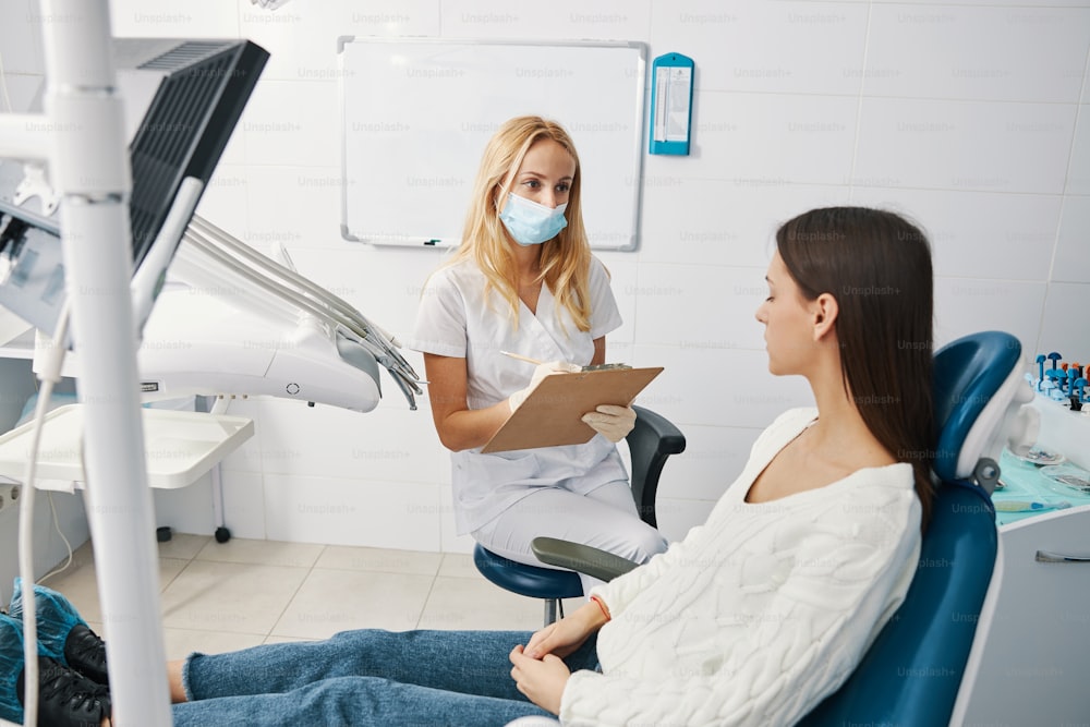 Mujer acostada en una silla dental con un dentista sentado cerca con un bloc de notas y un lápiz mientras escucha y toma notas dentro