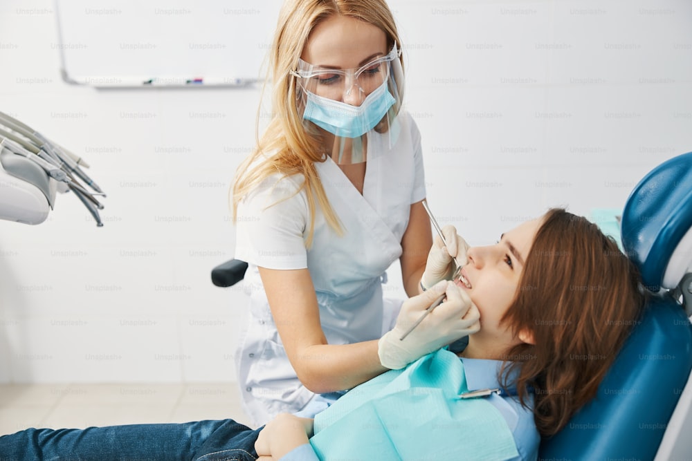 Cirujana dental femenina con escudo de plástico y máscara en la cara con sonda periodontal y espejo durante el chequeo de los dientes del niño
