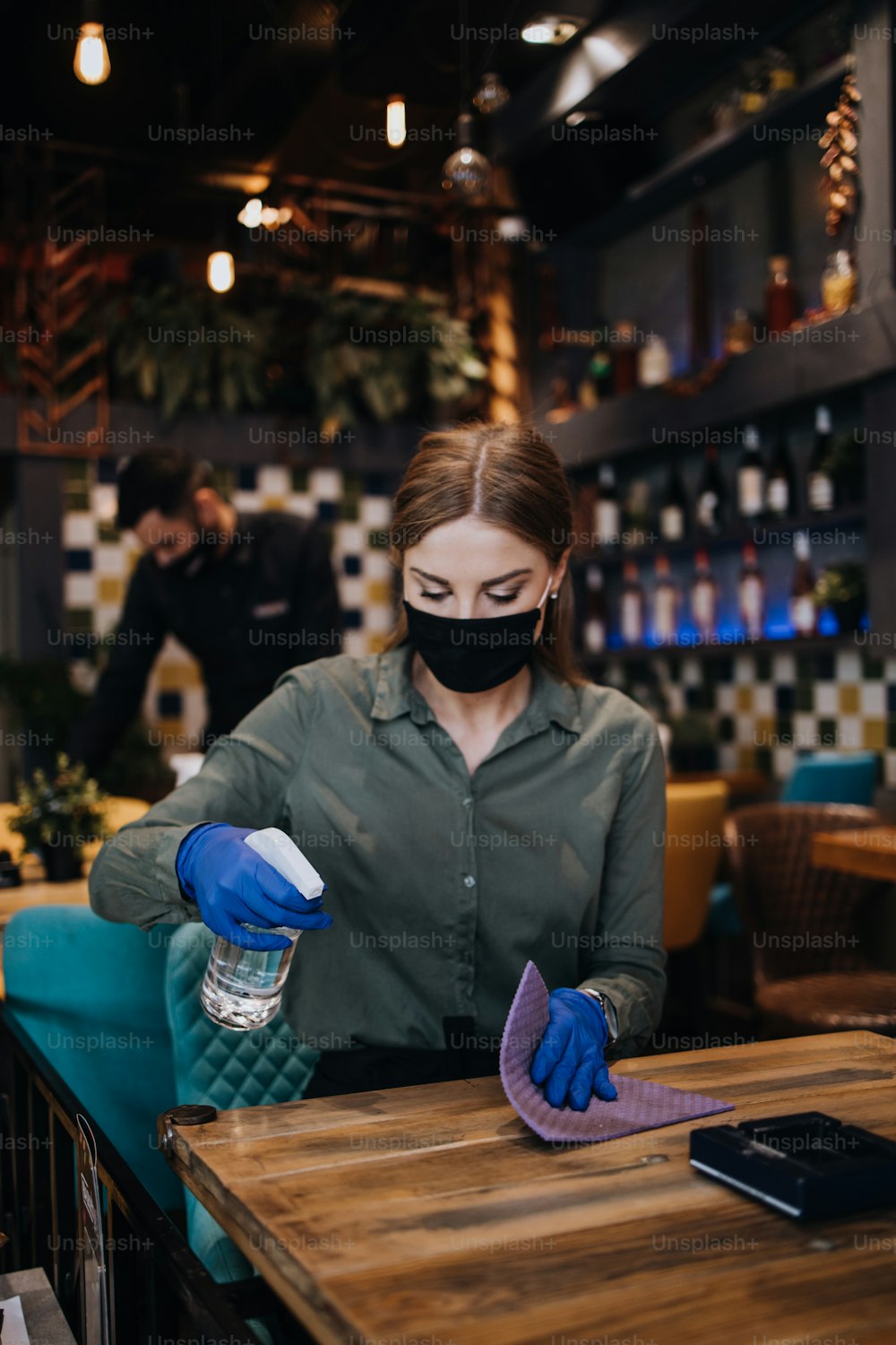 Young restaurant workers waiters cleaning and disinfecting tables and surfaces against Coronavirus pandemic disease. They are wearing protective face masks and gloves.