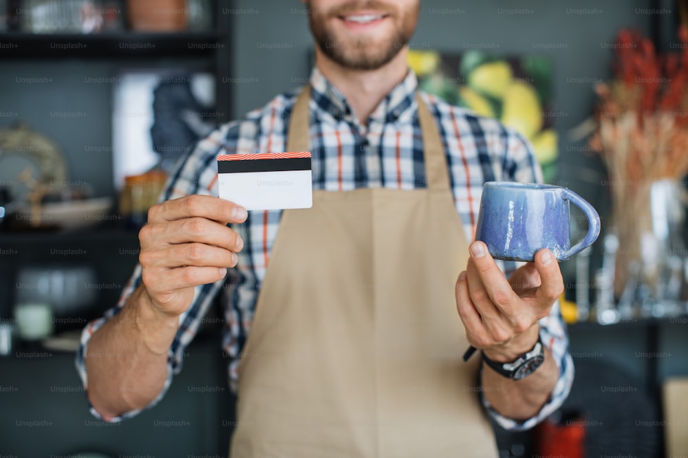 Positive bearded man holding ceramic cup and discount card in hands while posing at decor shop. Close up of hands of Caucasian salesman in beige apron showing credit card and blue cup to camera