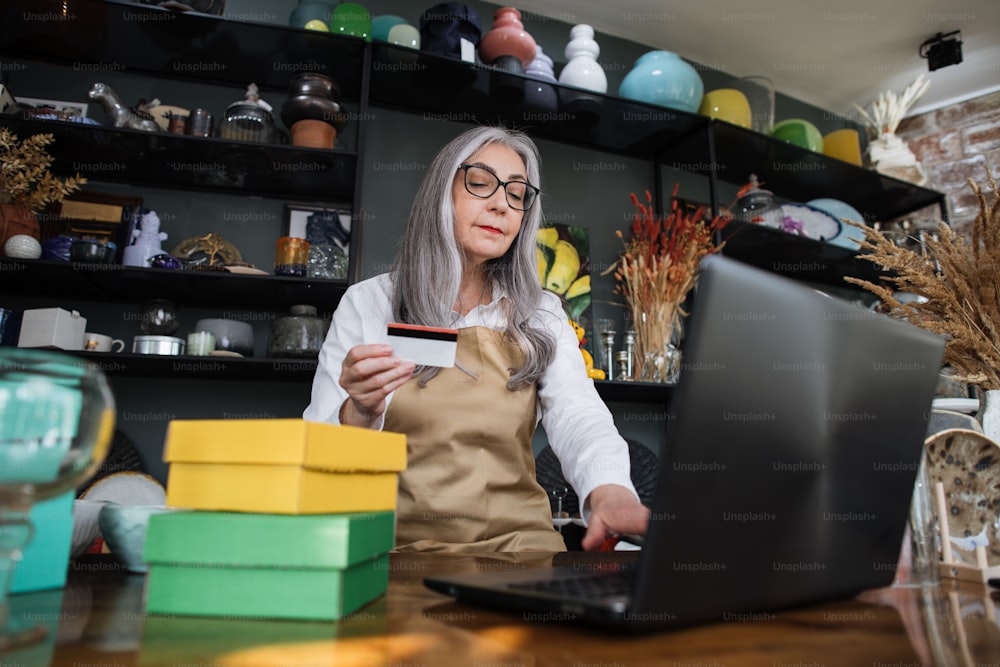 Shopping and discount system concept. Likable grey-haired lady typing on modern laptop while sitting at counter with credit discount card in hand. Shelves with various beautiful decor on background.
