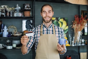 Positive bearded man holding ceramic cup and discount card in hands while posing at decor shop. Caucasian salesman in beige apron smiling and looking at camera.