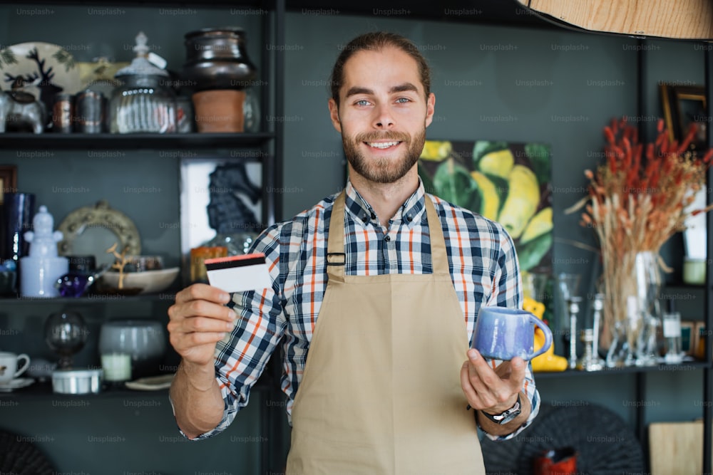 Positive bearded man holding ceramic cup and discount card in hands while posing at decor shop. Caucasian salesman in beige apron smiling and looking at camera.