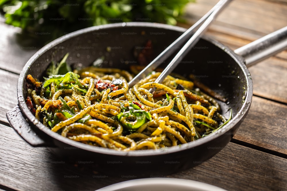 Spaghetti with pesto and parmesan cheese in a dark pan with tweezers in it.