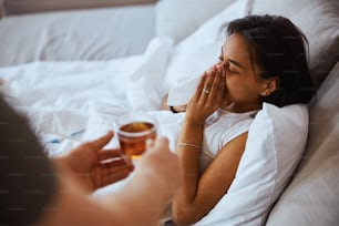 Primer plano de las manos masculinas sosteniendo una bebida caliente mientras una encantadora mujer joven acostada en la cama y sonándose la nariz en una servilleta de papel