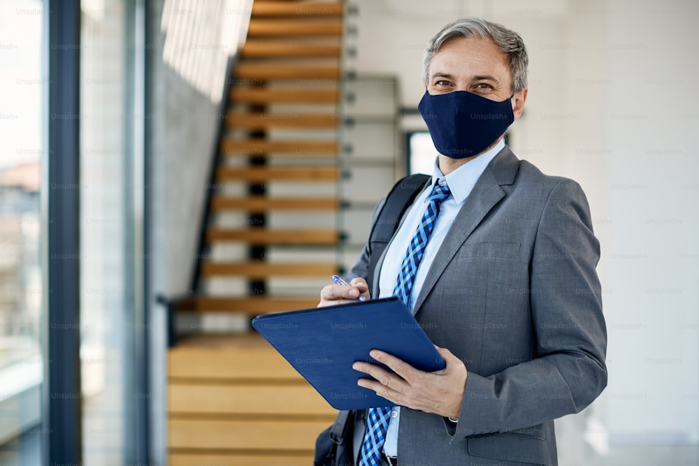 Happy real estate agent with face mask taking notes while checking apartment for sale and looking at camera.