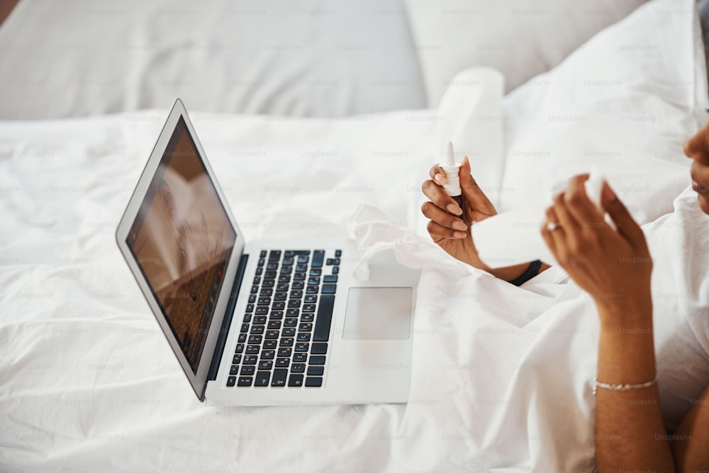 Gros plan d’une femme allongée dans son lit et tenant un spray nasal tout en utilisant un ordinateur portable et en parlant avec un médecin en ligne