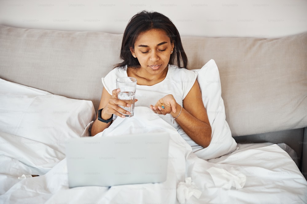 Hermosa dama afroamericana acostada en la cama con una computadora portátil y sonriendo mientras sostiene una píldora y un vaso de agua